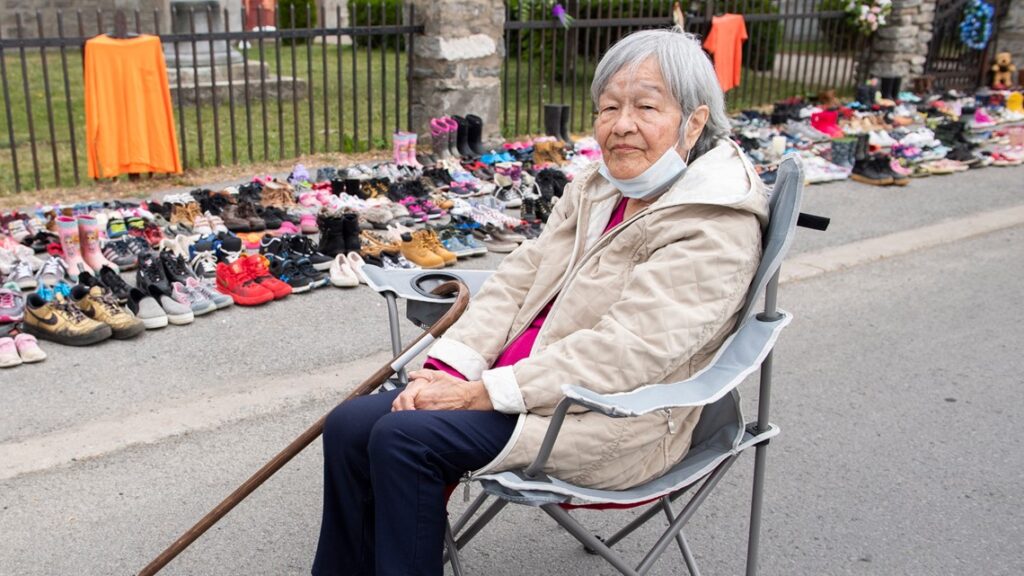 Residential school survivor Betty Deer sits next to children's shoes, placed there as a tribute to the victims of the residential school system outside St. Francis Xavier Church in Kahnawake, Quebec, Sunday, May 30, 2021.  Associated Press