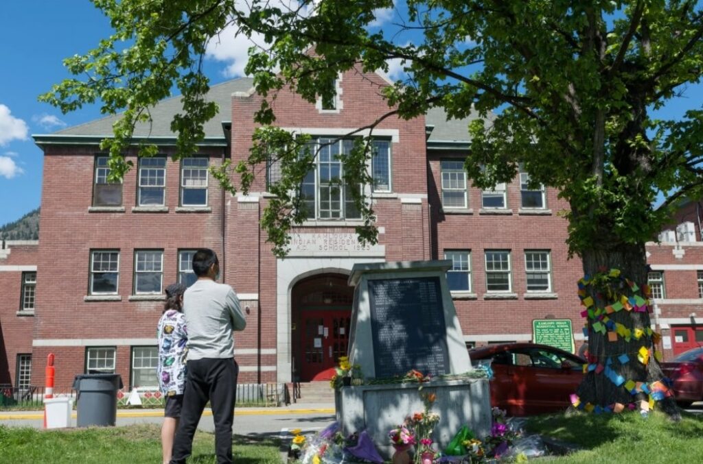 The remains were discovered during a search of the grounds of the former residential school, pictured above. Efforts are underway to recover the remains of the children. In this photo, a man stands with his son in front of a monument to the survivors of the former Kamloops Indian Residential School on May 29. (Dennis Owen / Reuters)