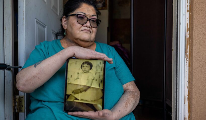 [Jacqueline Frost, 60, holds a photo showing how she was forced to adopt the look and attire of a white girl. She said she was beaten by a Ute aunt who served as a matron at a federal boarding school designed to assimilate Native children.Credit...Sharon Chischilly for The New York Times]