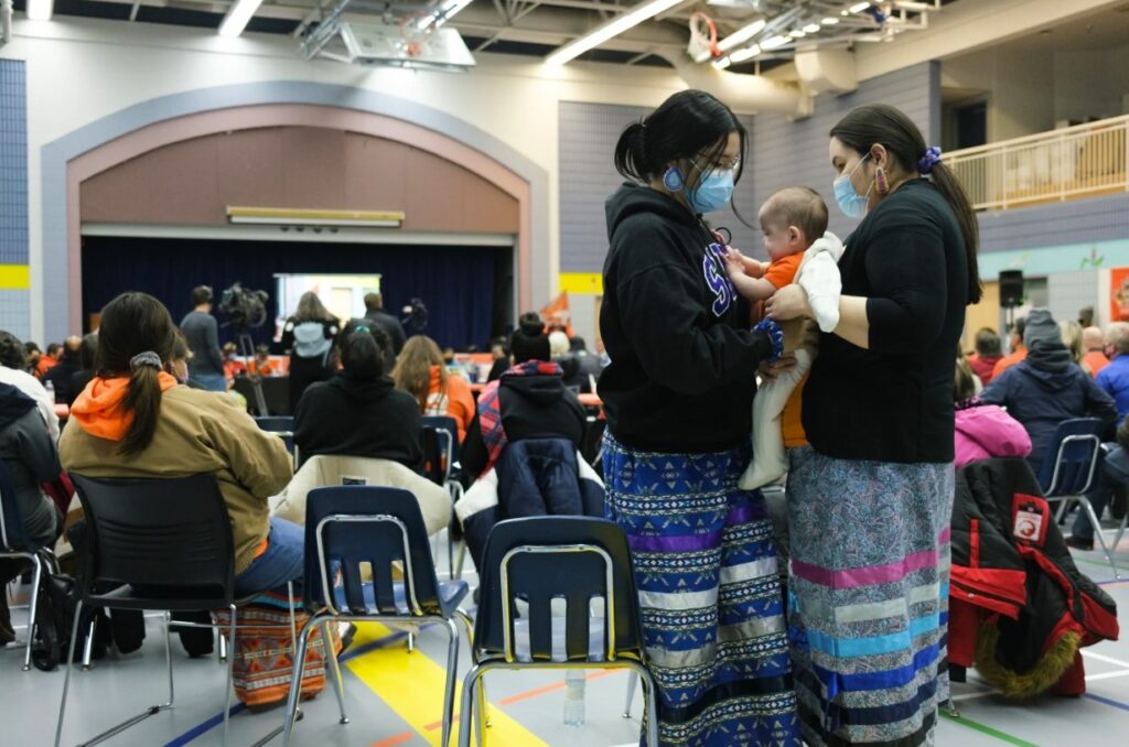 Pauline Pelly, far right, holds her daughter Ella during a public event at the Keeseekoose School Gymnasium at Keeseekoose First Nation on Tuesday Feb. 15, 2022. The event revealed that 54 graves had been found through ground-penetrating radar at the site of former residential schools St. Philip's and Fort Pelly. Michael Bell / The Canadian Press)