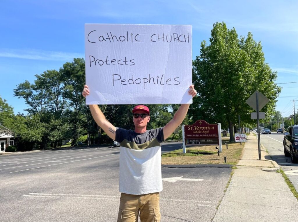 Ryan Brophy, holding one of several signs, is protesting this week near St. Veronica Catholic Chapel on Boston Neck Road. Photo by Amanda Milkovits