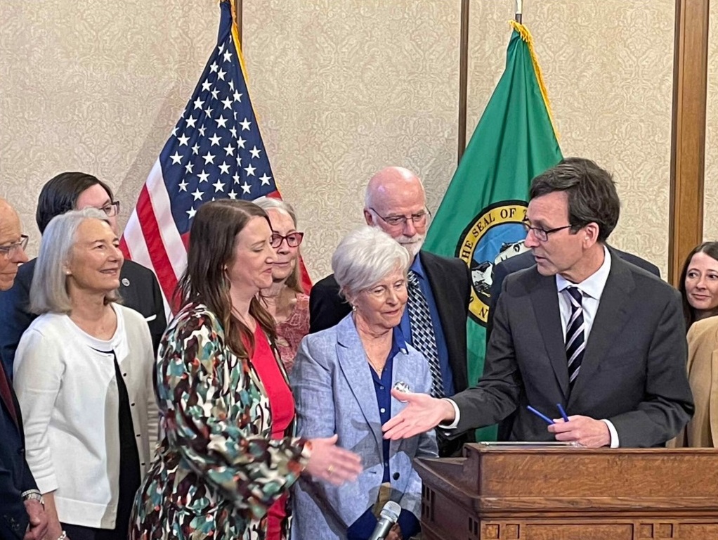 Gov. Bob Ferguson, at podium, goes to shake hands with state Sen. Noel Frame, D-Seattle, at the signing of a bill to make clergy mandatory reporters of child abuse and neglect, on May 2, 2025 in Olympia. Standing between them is Mary Dispenza, a founding member of the Catholic Accountability Project. (Photo by Jerry Cornfield/Washington State Standard)